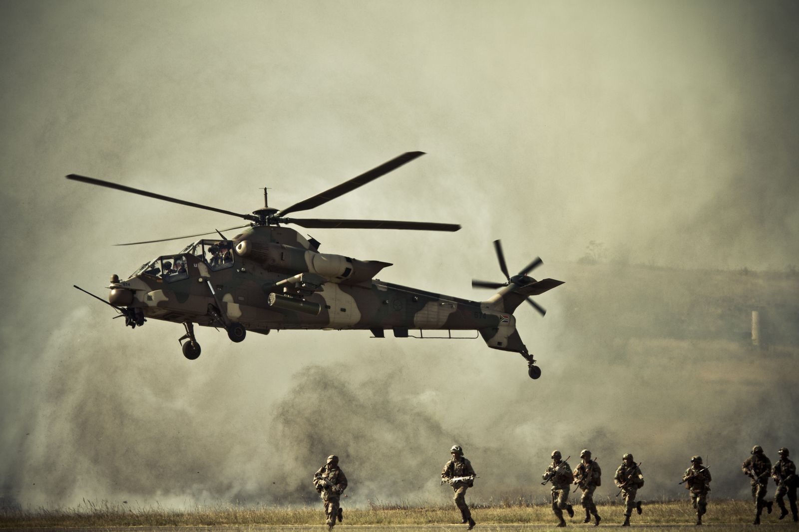 A military helicopter hovers as soldiers run on a misty battlefield.